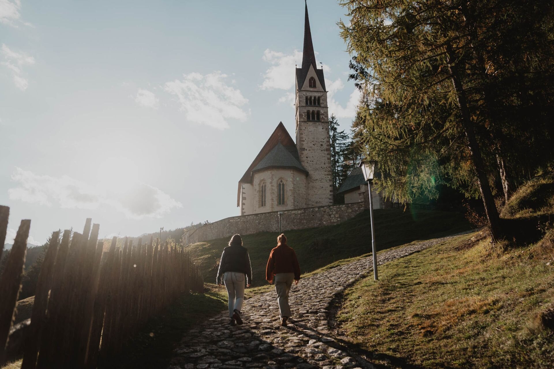 Due ragazze camminano verso una chiesetta nel bosco, tra i boschi autunnali della val di fassa | © Archivio immagini ApT Val di Fassa - Mattia Rizzi